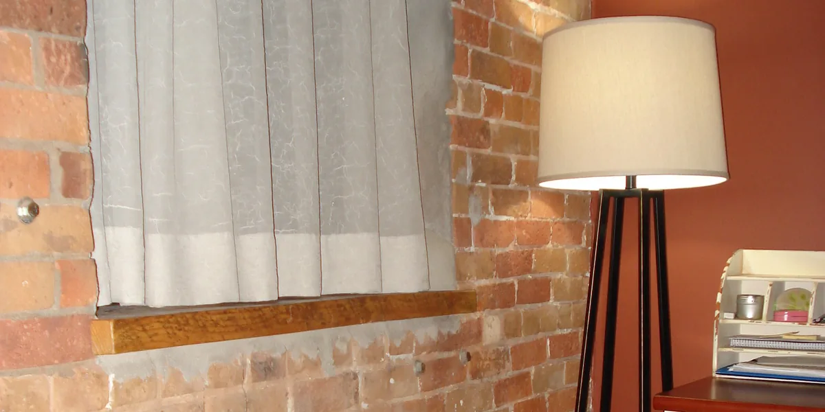 Stehlampe mit hellem Stoffschirm und schwarzem Gestell neben einem kleinen Schreibtisch mit Aufsatz, links ein Fenster mit Vorhang in einer Ziegelwand